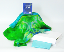 Load image into Gallery viewer, A_vibrant_green_and_blue_tie_dye_silk_scarf_draped_over_a_white_gift_box_with_a_light_blue_tissue_featuring_a_blue_MADE_LOCAL_card_on_top_set_against_a_white_background.
