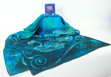 Load image into Gallery viewer, A_large_silk_shawl_draped_over_a_cube_in_vibrant_turquoise_and_dark_teal_greens_with_the_made_local_sign_displayed_in_front_of_a_white_background
